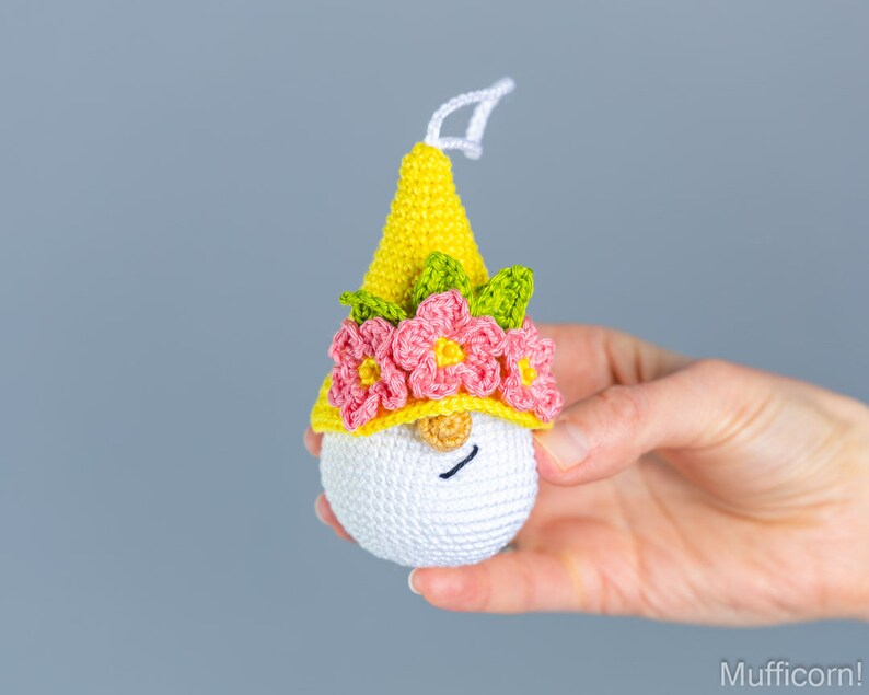 May include: A crocheted gnome ornament with a yellow hat, pink flowers, and green leaves. The gnome has a white face with a black smile.