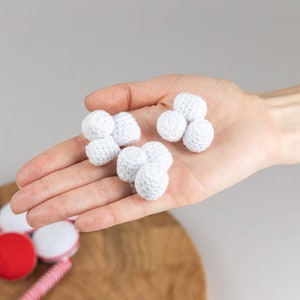 Puede incluir: Una mano que sostiene varias bolas peque&ntilde;as, blancas y tejidas a ganchillo. Las bolas est&aacute;n agrupadas en grupos de dos o tres. El fondo es de color gris claro. Otros objetos rojos y blancos son visibles en la parte inferior de la imagen.