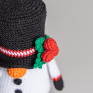 May include: A crocheted snowman ornament with a black top hat, red and green holly, and white and orange details.