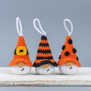 May include: Three handmade crochet gnome ornaments with orange and black hats, white bodies, and white hanging loops. The hats feature different designs: a yellow buckle, black stripes, and black polka dots. The gnomes have tan noses and smiling faces.