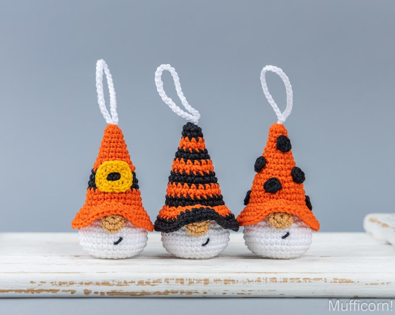 May include: Three handmade crochet gnome ornaments with orange and black hats, white bodies, and white hanging loops. The hats feature different designs: a yellow buckle, black stripes, and black polka dots. The gnomes have tan noses and smiling faces.