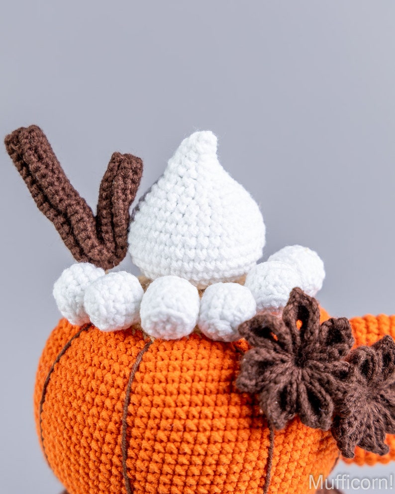 Puede incluir: Primer plano de una calabaza de ganchillo con la parte superior blanca y el tallo marr&oacute;n. La calabaza es naranja con l&iacute;neas verticales. Pompones blancos y una flor marr&oacute;n acent&uacute;an el dise&ntilde;o. La palabra "Mufficorn!" est&aacute; en la esquina inferior derecha.