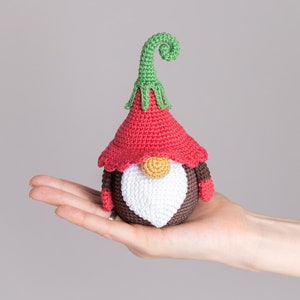 May include: A crocheted gnome with a red hat, white beard, and brown body. The gnome has a green curly hat top and is holding a small red mitten in each hand.
