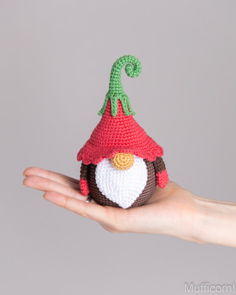 May include: A crocheted gnome with a red hat, white beard, and brown body. The gnome has a green curly hat top and is holding a small red mitten in each hand.