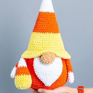 May include: A crocheted candy corn gnome with a white beard, orange and yellow hat, and a brown nose.