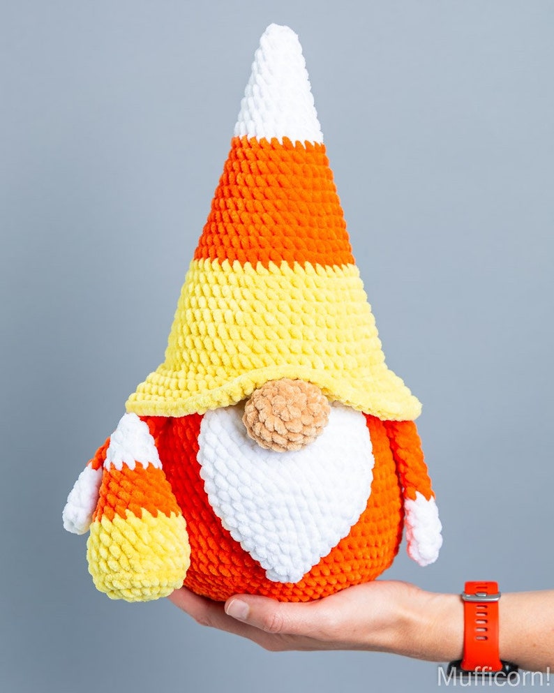 May include: A crocheted candy corn gnome with a white beard, orange and yellow hat, and a brown nose.
