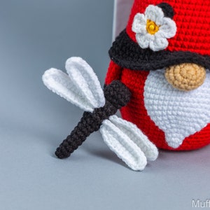 May include: A crocheted ladybug gnome with a white beard and a black hat with a white flower. The gnome is holding a crocheted dragonfly with black and white wings.