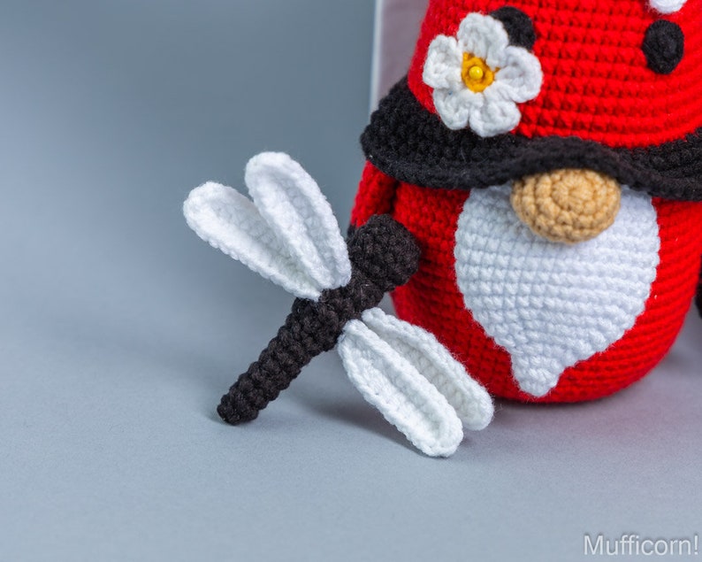 May include: A crocheted ladybug gnome with a white beard and a black hat with a white flower. The gnome is holding a crocheted dragonfly with black and white wings.