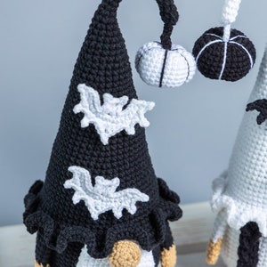 May include: Two crocheted gnomes dressed in black and white Halloween costumes. The gnome on the left has a black hat with white bats and a white pumpkin. The gnome on the right has a white hat with black bats and a black pumpkin.