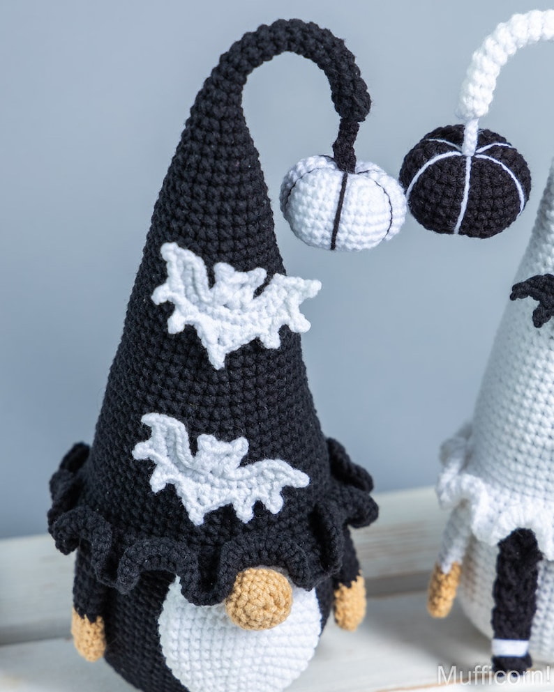 May include: Two crocheted gnomes dressed in black and white Halloween costumes. The gnome on the left has a black hat with white bats and a white pumpkin. The gnome on the right has a white hat with black bats and a black pumpkin.
