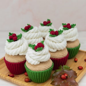 May include: Five crocheted cupcakes decorated with red berries and green leaves. The cupcakes are arranged on a wooden tray with a holly leaf and red berries.