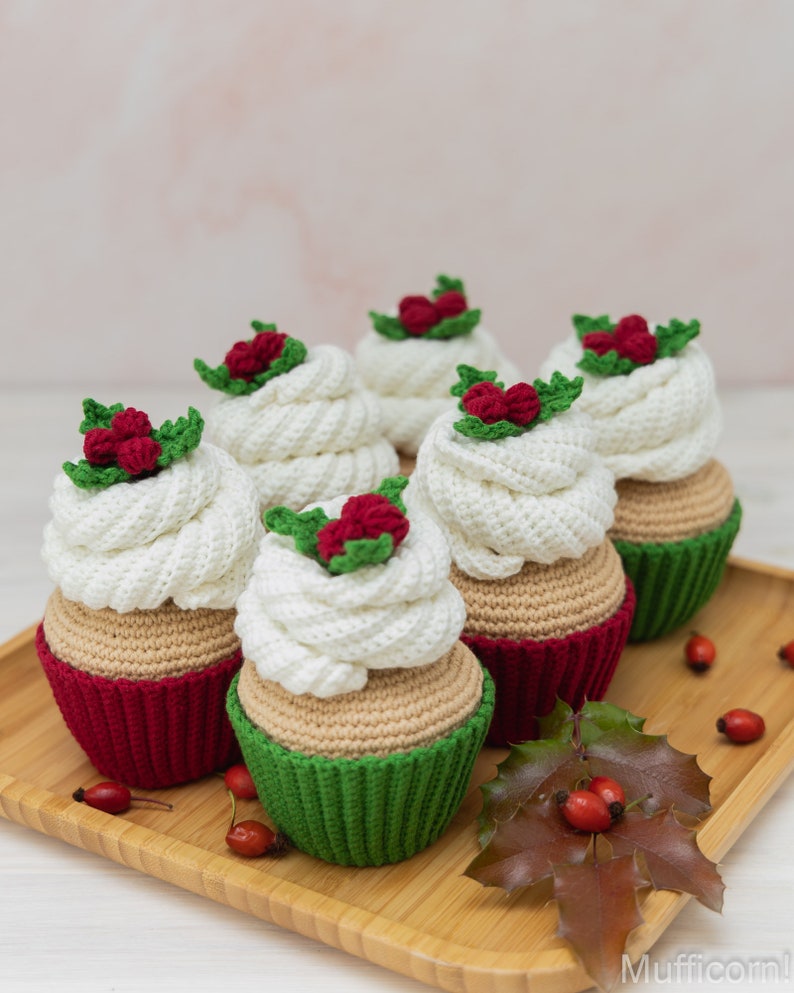 May include: Five crocheted cupcakes decorated with red berries and green leaves. The cupcakes are arranged on a wooden tray with a holly leaf and red berries.