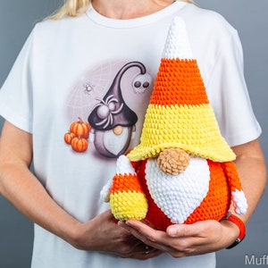May include: Two crocheted candy corn gnomes, one large and one small, are held in the hands of a person wearing a white t-shirt with a Halloween-themed design. The gnomes are made of orange, yellow, and white yarn.