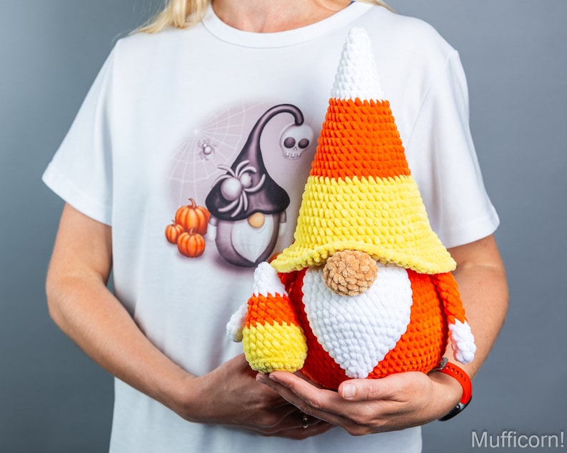 May include: Two crocheted candy corn gnomes, one large and one small, are held in the hands of a person wearing a white t-shirt with a Halloween-themed design. The gnomes are made of orange, yellow, and white yarn.