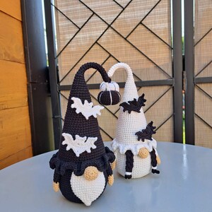 May include: Two crocheted gnomes, one black and one white, with white bat wings and a pumpkin on top of their hats. They are both wearing white beards and have black eyes.