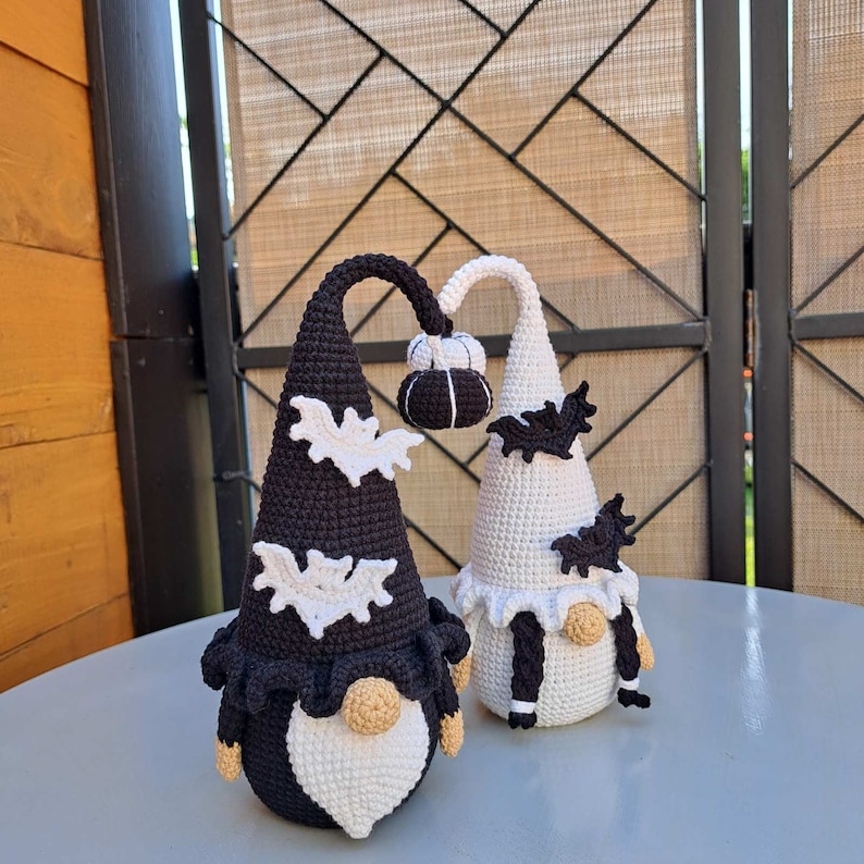 May include: Two crocheted gnomes, one black and one white, with white bat wings and a pumpkin on top of their hats. They are both wearing white beards and have black eyes.