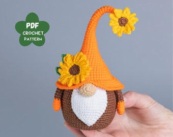 May include: A handmade crochet gnome with a brown body, white beard, and orange hat adorned with yellow sunflowers. The gnome has orange arms and a beige nose. The image includes a green flower-shaped graphic with the text "PDF CROCHET PATTERN".