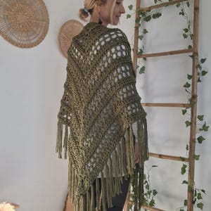 Op de afbeelding: Olijfgroene gehaakte sjaal met franjes. De sjaal wordt gedragen over een witte top en een zwarte broek. Het open weefpatroon is geometrisch. Het model draagt bruine laarzen en witte sokken. Een houten ladder en wanddecoratie zijn op de achtergrond.