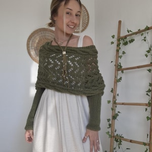 May include: An olive green knitted shawl with long sleeves and a diamond pattern. The shawl is worn over a white dress. A brown leather necklace with a tassel hangs from the neck. A wooden ladder with ivy is in the background.