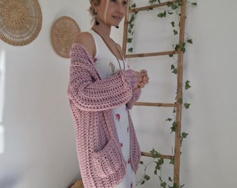 May include: A chunky knit cardigan in a soft dusty pink color featuring long sleeves, a relaxed open front, and two large patch pockets. The cardigan is worn over a sleeveless white floor-length dress decorated with a delicate red floral pattern.
