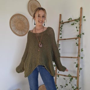 May include: An olive green, loose-knit, long-sleeved sweater with a wide neckline and a relaxed, oversized fit, paired with blue denim jeans that feature a flared leg cut. The sweater has a visible open-weave texture throughout the fabric.
