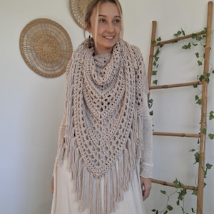 Peut inclure: Un châle en crochet beige à tissage ouvert avec des franges. Le châle est drapé sur les épaules d'une personne, associé à une longue robe crème. L'arrière-plan comprend des décorations murales et une échelle en bois ornée de verdure.