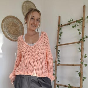 May include: A woman wearing a peach-colored, open-weave knit sweater with a deep V-neck. She is also wearing dark gray wide-leg pants. A wooden ladder with green vines is in the background, along with woven wall decor.