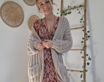 May include: A long-sleeved, chunky knit beige cardigan with two front pockets worn over a floor-length, V-neck dress featuring a maroon and white ikat-style geometric pattern with a tiered skirt and long sleeves.