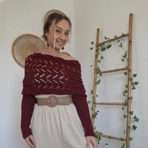 Könnte beinhalten: Ein weinroter Off-Shoulder-Pullover mit einem gemusterten Design. Der Pullover wird mit beigefarbenen Hosen mit weitem Bein und einem gewebten Gürtel mit einer kreisförmigen Schnalle kombiniert. Eine Bambusleiter mit Grünzeug befindet sich im Hintergrund.
