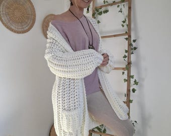 May include: A cream-colored, chunky knit cardigan with large pockets. The open-front sweater has a relaxed fit and long sleeves. The model is wearing a light purple top and light brown pants. The background includes decorative wall hangings and a wooden ladder.