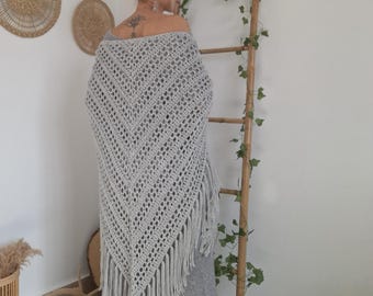 May include: A light gray, open-weave crochet shawl with fringe. The shawl is draped over the shoulders. The shawl is part of a bohemian style ensemble. The background includes decorative wall hangings and a wooden ladder.