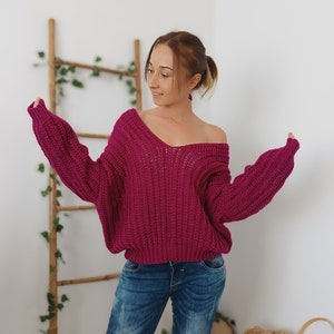 May include: A woman wearing a deep pink, oversized knitted sweater with a v-neck and long sleeves. She is wearing blue jeans.