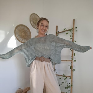 Pode incluir: Um suéter azul claro de malha aberta com mangas compridas e um cinto largo bege. O suéter é combinado com calças bege claro de pernas largas. A roupa é casual e confortável. O fundo inclui decorações de parede e uma escada de madeira.