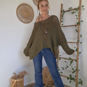May include: An olive green, loose-fitting, open-knit sweater with long sleeves and a wide neckline, paired with blue denim jeans. The sweater features a textured, chunky knit pattern that drapes over the shoulders and arms in a relaxed, oversized silhouette.