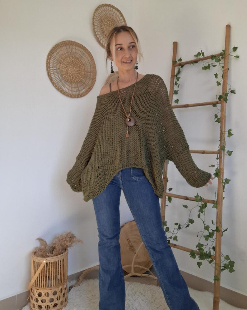 May include: An olive green, loose-fitting, open-knit sweater with long sleeves and a wide neckline, paired with blue denim jeans. The sweater features a textured, chunky knit pattern that drapes over the shoulders and arms in a relaxed, oversized silhouette.
