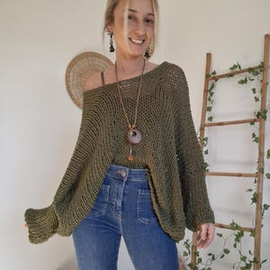 May include: An olive green, loose-knit sweater with a wide, open-weave texture, long sleeves, and a relaxed, oversized fit. The garment features a wide neckline and is paired with blue denim jeans and a long pendant necklace with a circular charm.