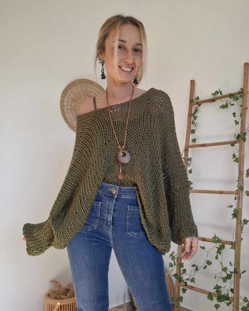 May include: An olive green, loose-knit sweater with a wide, open-weave texture, long sleeves, and a relaxed, oversized fit. The garment features a wide neckline and is paired with blue denim jeans and a long pendant necklace with a circular charm.