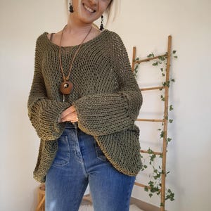 May include: An olive green, loose-knit sweater with a wide neckline and long, cuffed sleeves, paired with blue denim jeans featuring front patch pockets. The sweater has a chunky, open-weave texture and a relaxed, oversized fit throughout the body.