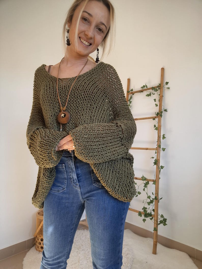 May include: An olive green, loose-knit sweater with a wide neckline and long, cuffed sleeves, paired with blue denim jeans featuring front patch pockets. The sweater has a chunky, open-weave texture and a relaxed, oversized fit throughout the body.