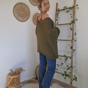 May include: An olive green, loose-knit, oversized sweater with a wide neckline and long sleeves, paired with blue denim jeans. The sweater features a chunky, open-weave texture and a relaxed fit that drapes over the shoulders and torso.