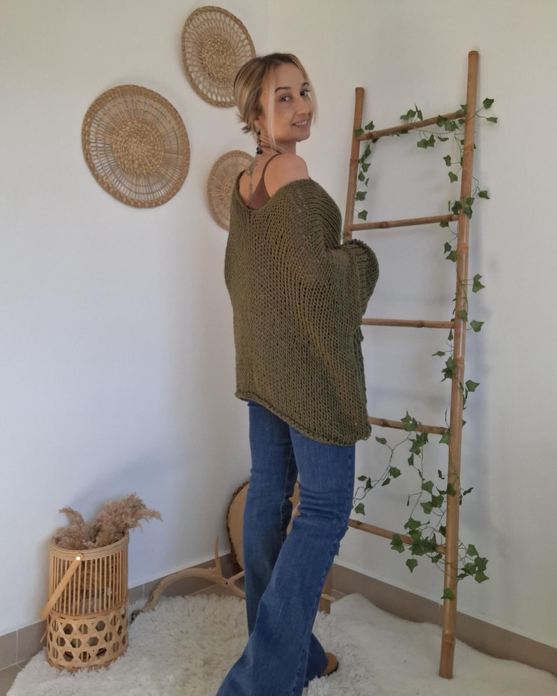May include: An olive green, loose-knit, oversized sweater with a wide neckline and long sleeves, paired with blue denim jeans. The sweater features a chunky, open-weave texture and a relaxed fit that drapes over the shoulders and torso.