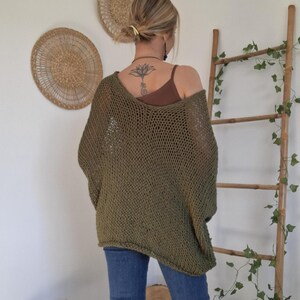 May include: An olive green, loose-fitting, open-knit sweater with a wide neckline and long sleeves. The garment features a coarse, textured weave pattern and is worn over a brown tank top, paired with blue denim jeans.
