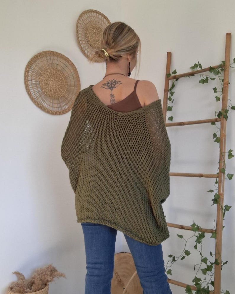 May include: An olive green, loose-fitting, open-knit sweater with a wide neckline and long sleeves. The garment features a coarse, textured weave pattern and is worn over a brown tank top, paired with blue denim jeans.