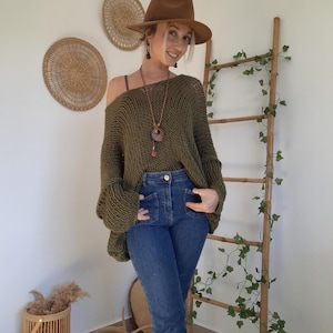 May include: An olive green loose-knit sweater with a wide neckline, paired with blue denim jeans, a brown fedora hat, and a long necklace featuring a circular wooden pendant. The sweater has long sleeves and a textured, open-weave construction.