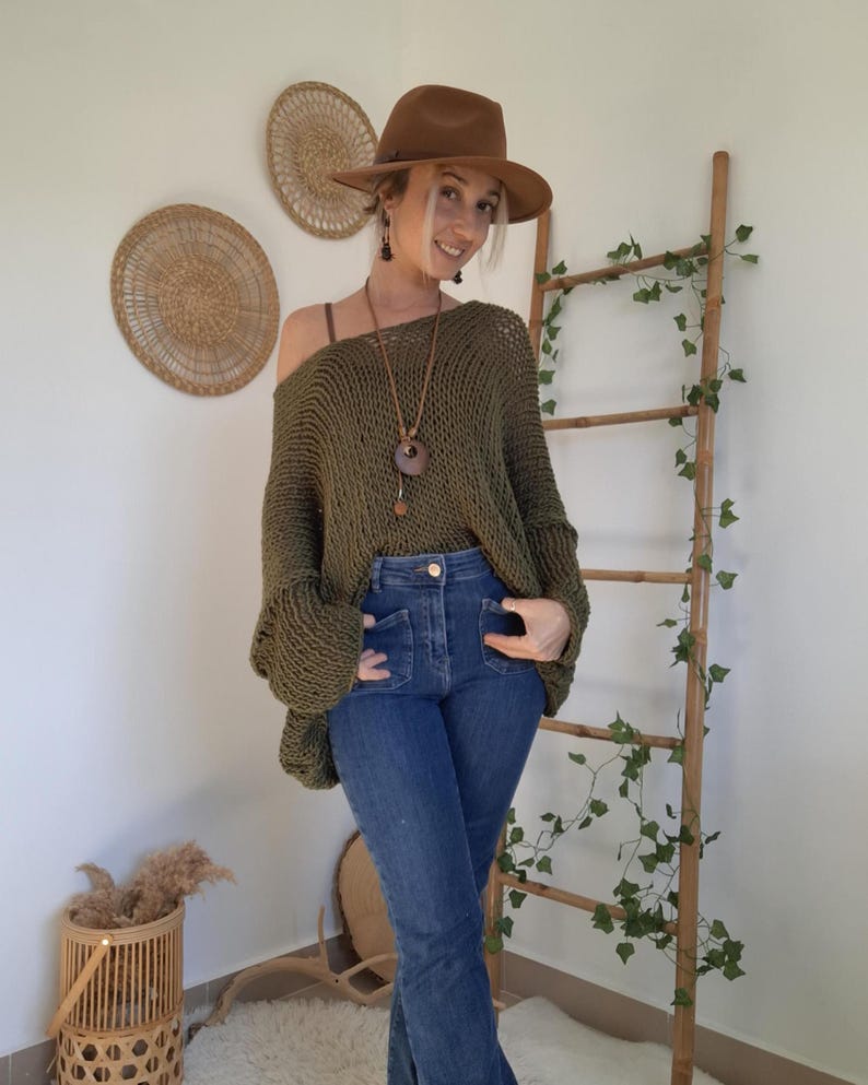May include: An olive green loose-knit sweater with a wide neckline, paired with blue denim jeans, a brown fedora hat, and a long necklace featuring a circular wooden pendant. The sweater has long sleeves and a textured, open-weave construction.
