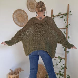 May include: An olive green, loose-fitting, open-knit sweater with long, wide sleeves and a wide neckline. The garment features a textured, net-like stitch pattern throughout, providing a relaxed, oversized silhouette that drapes over the shoulders.