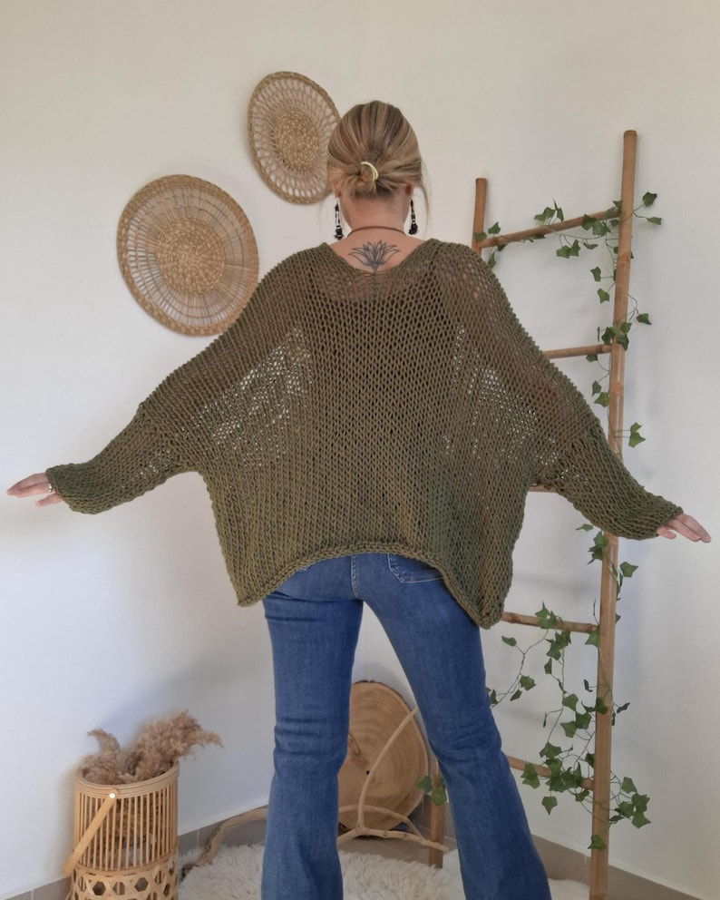 May include: An olive green, loose-fitting, open-knit sweater with long, wide sleeves and a wide neckline. The garment features a textured, net-like stitch pattern throughout, providing a relaxed, oversized silhouette that drapes over the shoulders.
