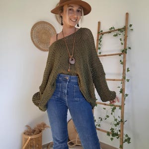 May include: An olive green loose-knit sweater with a wide neckline, paired with blue denim jeans featuring front patch pockets, a brown fedora hat, and a long necklace with a circular pendant. The sweater has long sleeves and a relaxed fit.