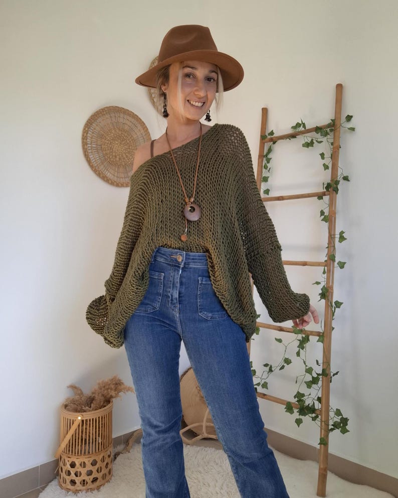 May include: An olive green loose-knit sweater with a wide neckline, paired with blue denim jeans featuring front patch pockets, a brown fedora hat, and a long necklace with a circular pendant. The sweater has long sleeves and a relaxed fit.