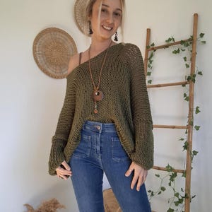 May include: An olive green, loose-knit, long-sleeved sweater with a wide neckline, paired with blue denim jeans featuring front patch pockets. A long brown leather cord necklace with a circular wooden pendant hangs around the neck.